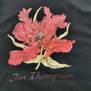 May include: Black t-shirt featuring a large, detailed red flower print with green and yellow accents. The text "Jim Thompson" is printed in a brown font at the bottom of the shirt. The floral design is centered on the shirt.