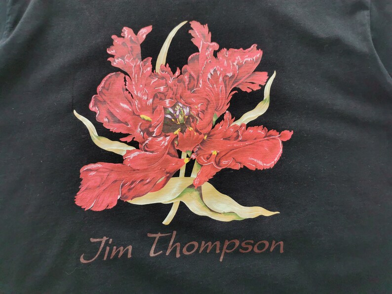 May include: Black t-shirt featuring a large, detailed red flower print with green and yellow accents. The text "Jim Thompson" is printed in a brown font at the bottom of the shirt. The floral design is centered on the shirt.
