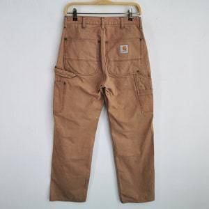 May include: Tan work pants with multiple pockets and a Carhartt logo patch on the back. The pants are hanging on a wooden hanger against a white wall. The pants have a straight leg and a classic workwear design.