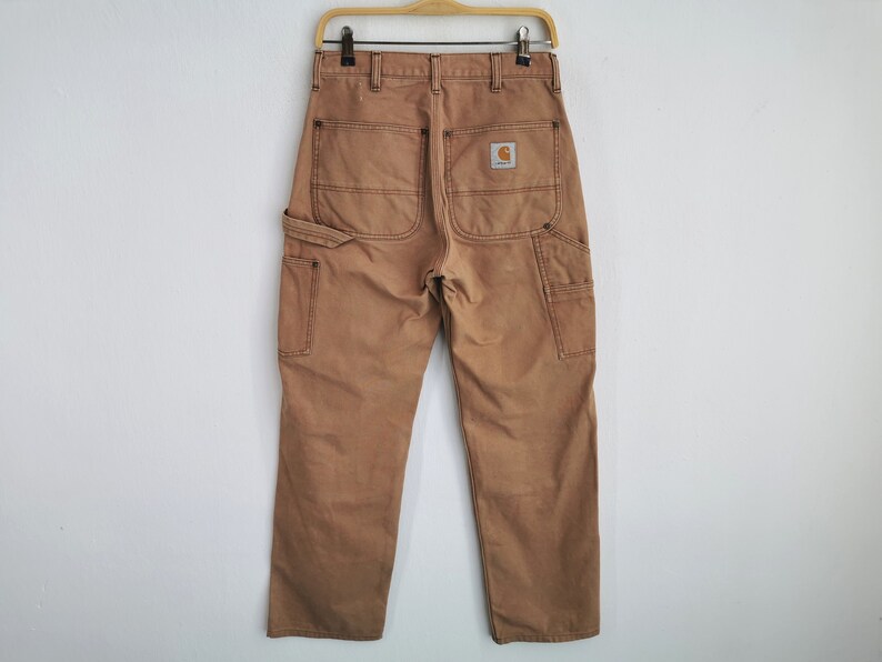 May include: Tan work pants with multiple pockets and a Carhartt logo patch on the back. The pants are hanging on a wooden hanger against a white wall. The pants have a straight leg and a classic workwear design.