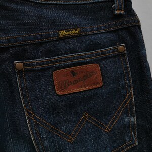 May include: Close-up of dark blue denim jeans with the Wrangler logo on the back pocket. The jeans feature a brown leather patch with the Wrangler logo and stitching details. The stitching is in a contrasting orange colour.