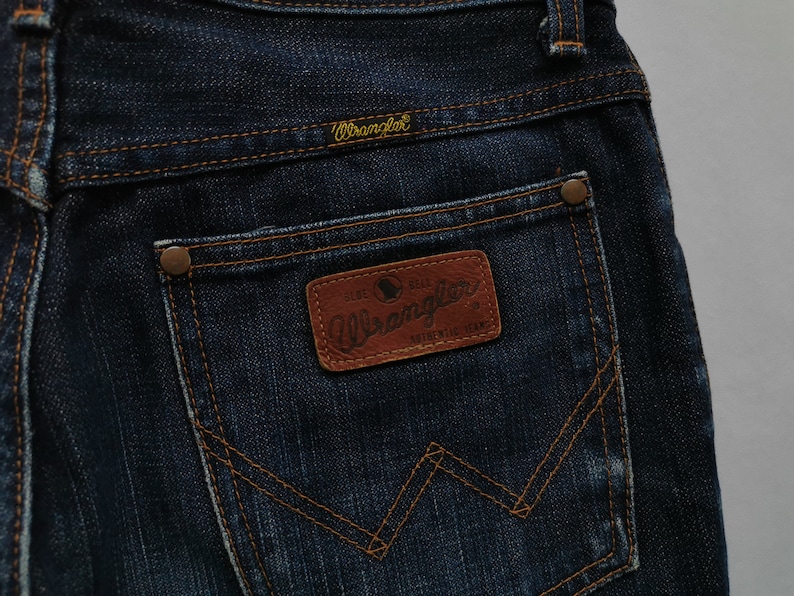 May include: Close-up of dark blue denim jeans with the Wrangler logo on the back pocket. The jeans feature a brown leather patch with the Wrangler logo and stitching details. The stitching is in a contrasting orange colour.