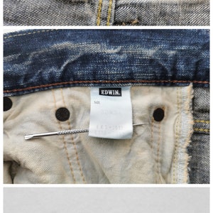 May include: Close-up of blue denim jeans with visible labels. The labels display the brand "EDWIN", size 32x34, and "MADE IN JAPAN". The jeans have a classic five-pocket design and a button fly.