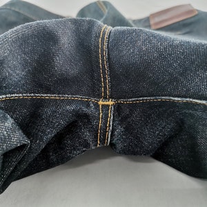 May include: Close-up of dark blue denim jeans. The jeans feature visible stitching in a contrasting yellow-orange thread. A brown leather patch is visible at the top. The fabric has a textured appearance.