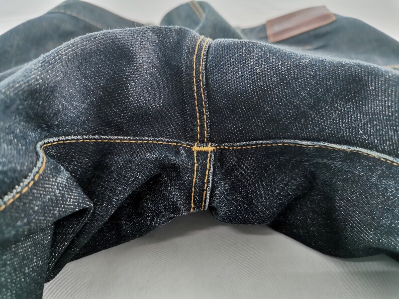 May include: Close-up of dark blue denim jeans. The jeans feature visible stitching in a contrasting yellow-orange thread. A brown leather patch is visible at the top. The fabric has a textured appearance.