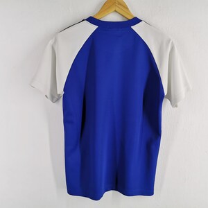 Puede incluir: Camiseta de manga corta azul y blanca colgada de una percha de madera. La camiseta tiene un cuerpo azul y mangas blancas. Tiene cuello redondo y una raya negra en la nuca. Sobre una pared blanca.