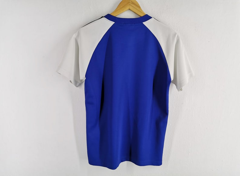 Puede incluir: Camiseta de manga corta azul y blanca colgada de una percha de madera. La camiseta tiene un cuerpo azul y mangas blancas. Tiene cuello redondo y una raya negra en la nuca. Sobre una pared blanca.