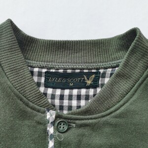 May include: Close-up of a green sweatshirt with a ribbed collar and buttoned placket. The interior collar features a black and white gingham pattern. A label reads "LYLE & SCOTT" with a golden eagle emblem.