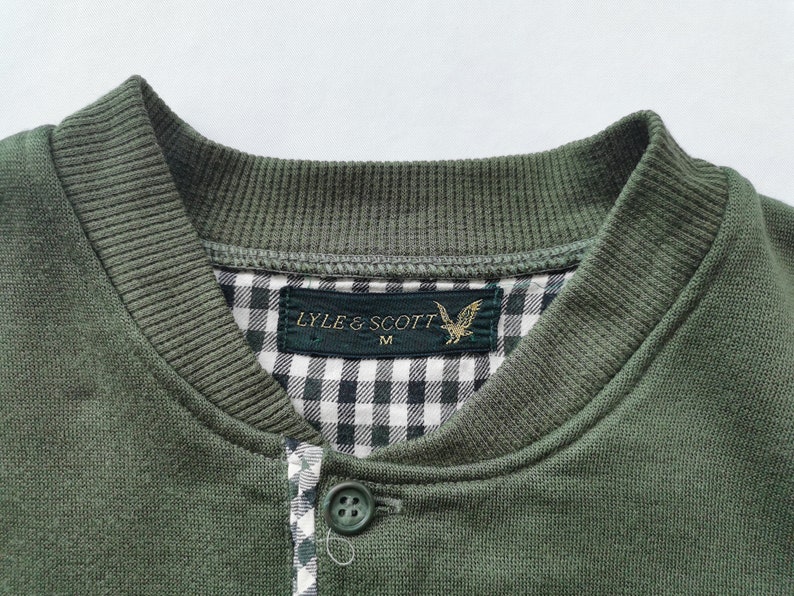 May include: Close-up of a green sweatshirt with a ribbed collar and buttoned placket. The interior collar features a black and white gingham pattern. A label reads "LYLE & SCOTT" with a golden eagle emblem.