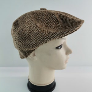Op de afbeelding: Bruin en beige newsboy-pet met visgraatpatroon. De pet wordt gepresenteerd op een mannequinhoofd tegen een witte achtergrond. Hij heeft een ronde kroon en een kleine, stijve klep.