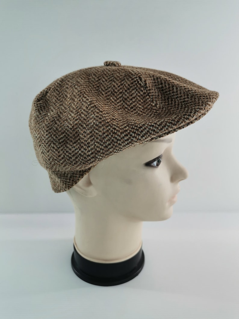Op de afbeelding: Bruin en beige newsboy-pet met visgraatpatroon. De pet wordt gepresenteerd op een mannequinhoofd tegen een witte achtergrond. Hij heeft een ronde kroon en een kleine, stijve klep.