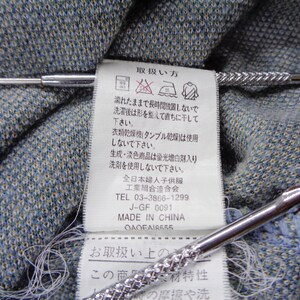Puede incluir: Primer plano de una tela azul y gris texturizada con una etiqueta blanca con texto japon&eacute;s y s&iacute;mbolos de cuidado. Dos alfileres de corbata plateados con dise&ntilde;os texturizados est&aacute;n colocados sobre la tela.
