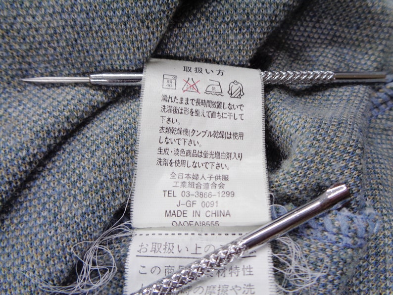 Puede incluir: Primer plano de una tela azul y gris texturizada con una etiqueta blanca con texto japon&eacute;s y s&iacute;mbolos de cuidado. Dos alfileres de corbata plateados con dise&ntilde;os texturizados est&aacute;n colocados sobre la tela.
