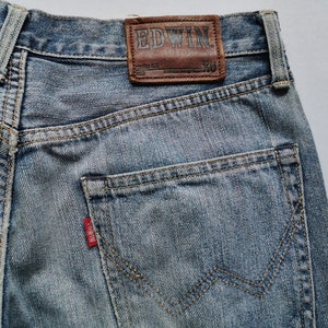 May include: Close-up of light blue denim jeans. The jeans have a brown leather patch with the brand name "EDWIN" and a red tag on the back pocket. The jeans feature a classic five-pocket design and are made of durable denim.