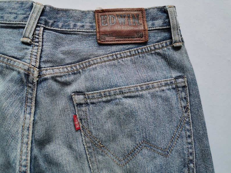 May include: Close-up of light blue denim jeans. The jeans have a brown leather patch with the brand name "EDWIN" and a red tag on the back pocket. The jeans feature a classic five-pocket design and are made of durable denim.