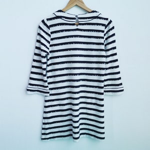 Puede incluir: Vestido a rayas blancas y negras con mangas tres cuartos colgado de una percha de madera. El vestido presenta un cuello redondo y un detalle de ojo de cerradura en la espalda. Las rayas son horizontales y espaciadas uniformemente.