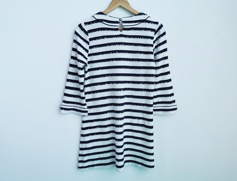 Puede incluir: Vestido a rayas blancas y negras con mangas tres cuartos colgado de una percha de madera. El vestido presenta un cuello redondo y un detalle de ojo de cerradura en la espalda. Las rayas son horizontales y espaciadas uniformemente.