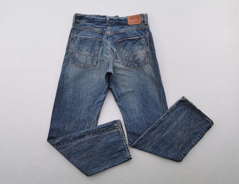May include: Blue denim jeans with a faded wash and classic five-pocket design. The jeans have a straight leg cut and a leather patch on the back waistband. The denim has a worn look.
