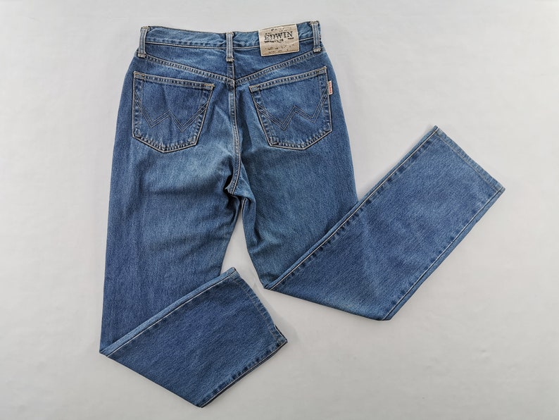 Puede incluir: Vaqueros de mezclilla azul con un dise&ntilde;o cl&aacute;sico de cinco bolsillos. Los vaqueros tienen un corte de pierna recta y un lavado descolorido. Los bolsillos traseros tienen un patr&oacute;n de costura distintivo. Una peque&ntilde;a etiqueta con la marca "EDWIN" es visible.