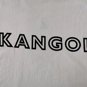 Op de afbeelding: Witte stof met het zwarte woord "KANGOL" in het midden gedrukt. De tekst is in een vette, schreefloze lettertype met een dubbele omtrek. De stof heeft een licht gekreukte textuur.