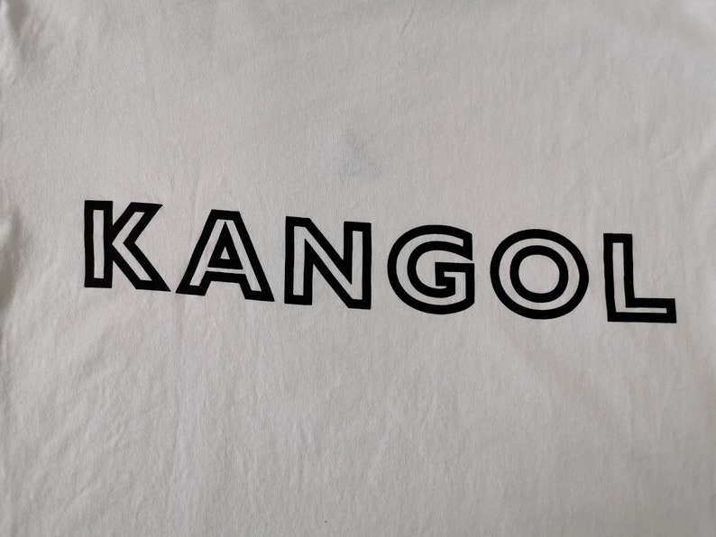 Op de afbeelding: Witte stof met het zwarte woord "KANGOL" in het midden gedrukt. De tekst is in een vette, schreefloze lettertype met een dubbele omtrek. De stof heeft een licht gekreukte textuur.