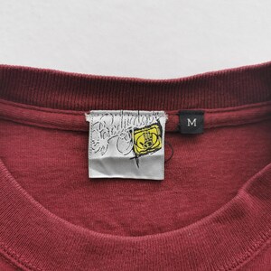 May include: A close-up shot of a maroon crew neck t-shirt. The shirt features a brand label with a yellow hand graphic. A black label with the letter "M" is also visible. The garment is made of a soft material.
