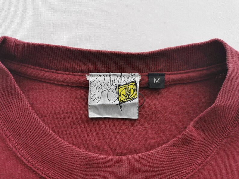 May include: A close-up shot of a maroon crew neck t-shirt. The shirt features a brand label with a yellow hand graphic. A black label with the letter "M" is also visible. The garment is made of a soft material.
