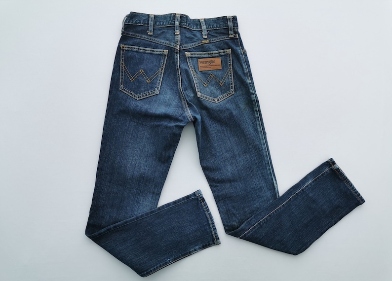 May include: Dark blue denim jeans with a classic five-pocket design. Features a brown leather patch with the "Wrangler" logo on the back. The jeans have a straight leg cut and are laid flat on a white surface.