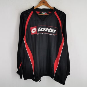 Puede incluir: Camiseta deportiva negra de manga larga con detalles rojos y el logo Lotto. La camiseta tiene cuello en V y un cordón en la parte inferior. El logo Lotto es blanco con borde rojo y el texto "Italian Sport Design".