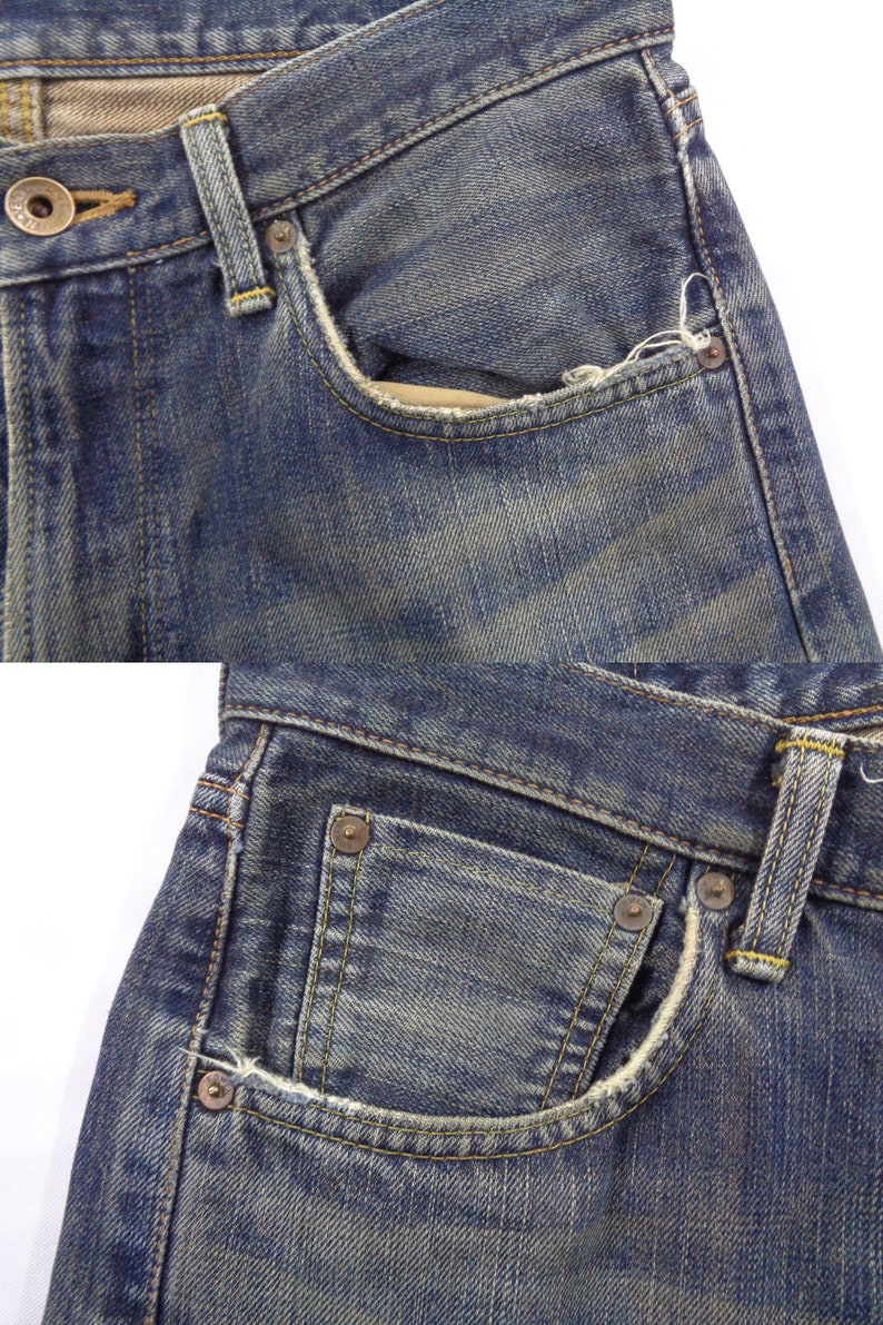 Puede incluir: Primer plano de unos vaqueros de mezclilla azules, que muestran los bolsillos delanteros, el bot&oacute;n y las trabillas. Los vaqueros tienen un aspecto desgastado y descolorido con costuras y remaches met&aacute;licos visibles. La tela est&aacute; desgastada, con algunos deshilachados.