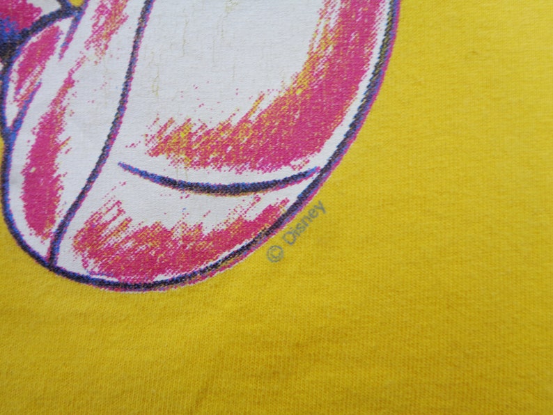 Op de afbeelding: Close-up van gele stof met een grafisch ontwerp in wit, roze en blauw. Het ontwerp is een gedeeltelijke afbeelding van een personage. Het Disney copyright symbool is zichtbaar.