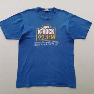 Può includere: T-shirt blu con il logo K-ROCK 92.3 FM in un riquadro giallo e bianco, con una grafica di chitarra. Il testo "Howard Stern All Morning Classic Rock & Roll All Day" è stampato sotto.