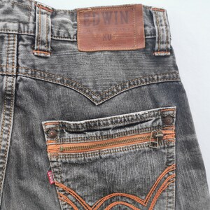May include: Close-up of the back of gray denim jeans with orange stitching and zipper details. The jeans feature a brown leather patch with the brand name "EDWIN" and "EXCLUSIVE VINTAGE" text. The back pocket has a zipper closure.