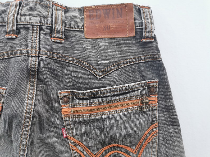 May include: Close-up of the back of gray denim jeans with orange stitching and zipper details. The jeans feature a brown leather patch with the brand name "EDWIN" and "EXCLUSIVE VINTAGE" text. The back pocket has a zipper closure.