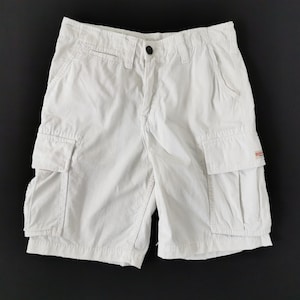 May include: Cream-coloured cargo shorts featuring multiple pockets and a button fastening. The shorts have a classic design and are made of a durable fabric. The shorts are displayed on a black background.