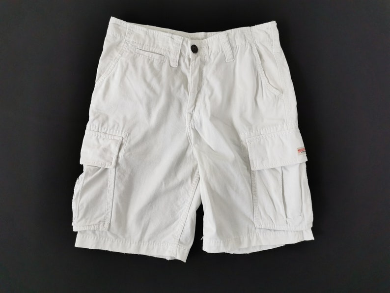 May include: Cream-coloured cargo shorts featuring multiple pockets and a button fastening. The shorts have a classic design and are made of a durable fabric. The shorts are displayed on a black background.