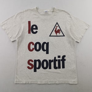 May include: A light grey t-shirt featuring the text "le coq sportif" in red and navy blue. The brand's logo, a red rooster within a black triangle, is also visible. The shirt has short sleeves and a crew neck.