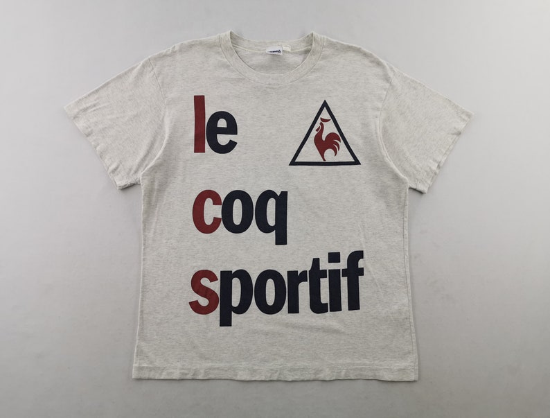 May include: A light grey t-shirt featuring the text "le coq sportif" in red and navy blue. The brand's logo, a red rooster within a black triangle, is also visible. The shirt has short sleeves and a crew neck.