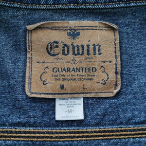 May include: Close-up of a denim jacket with a leather label. The label reads "Edwin Tokyo Japan Guaranteed" with additional text. The jacket has a blue hue with visible stitching and a smaller tag below the main label.