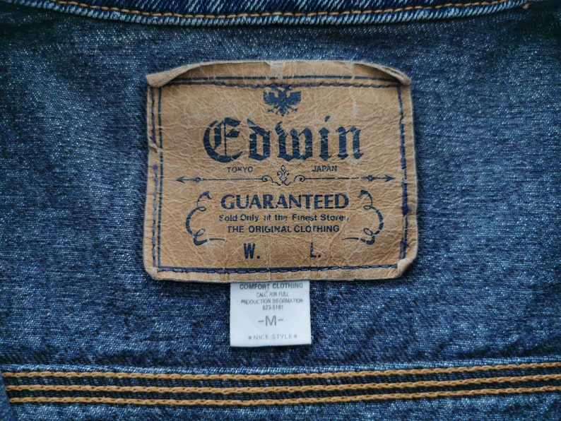 May include: Close-up of a denim jacket with a leather label. The label reads "Edwin Tokyo Japan Guaranteed" with additional text. The jacket has a blue hue with visible stitching and a smaller tag below the main label.