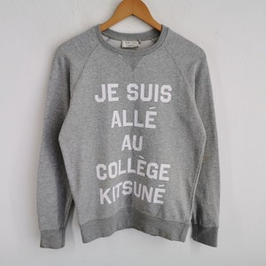 May include: A heather gray sweatshirt with the text "JE SUIS ALLÉ AU COLLÈGE KITSUNÉ" in white block letters. The sweatshirt has a crew neck and long sleeves, and is hanging on a wooden hanger.