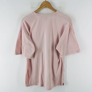 May include: A light pink t-shirt with short sleeves, hanging on a wooden hanger. The shirt is made of a soft material and has a relaxed fit. The background is a plain white wall.