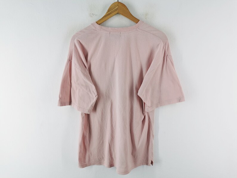 May include: A light pink t-shirt with short sleeves, hanging on a wooden hanger. The shirt is made of a soft material and has a relaxed fit. The background is a plain white wall.