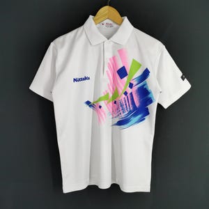 Puede incluir: Polo blanco con un diseño abstracto colorido en rosa, azul y verde. La camisa presenta el nombre de la marca "Nittaku" en azul en el pecho. Manga corta, con cuello.