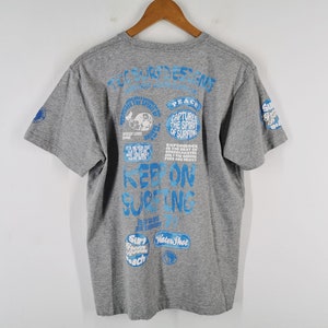 May include: Gray t-shirt with blue text and graphics. The back of the shirt features various phrases and images related to surfing, including "Keep on Surfing" and "Captures the Spirit of Surfing."