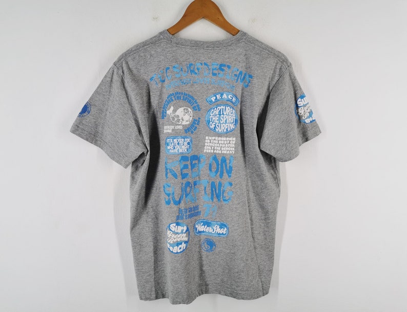 May include: Gray t-shirt with blue text and graphics. The back of the shirt features various phrases and images related to surfing, including "Keep on Surfing" and "Captures the Spirit of Surfing."