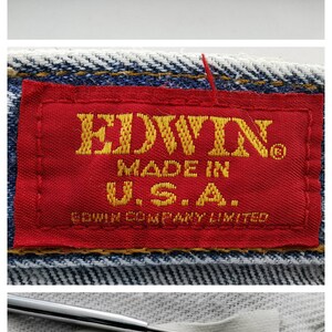 Może przedstawiać: Zbliżenie jasnoniebieskich dżins&oacute;w. Obraz przedstawia tylny szew, czerwoną etykietę z napisem "EDWIN MADE IN U.S.A." oraz białą metkę z rozmiarem 34x32. Dżinsy są wykonane w 100% z bawełny.