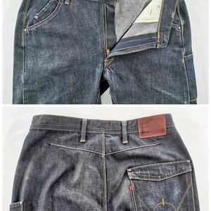 May include: Dark wash denim jeans with a button and zip fastening. Features include front pockets with rivets, a coin pocket, and a back pocket with a distinctive stitch design. The brand name "E FUNCTION" is visible on a red tag.