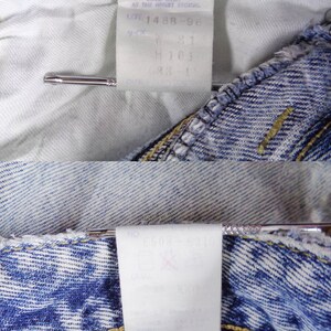 May include: Close-up of the inside of blue denim jeans, showing the white fabric and tags. The tags display text including size information and care instructions. A silver metal pin is holding the fabric together.