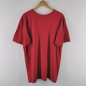 May include: A solid red t-shirt with a crew neck and short sleeves hangs from a wooden hanger. The shirt is made of a soft material and is displayed against a plain white background. The t-shirt is a basic, casual garment.